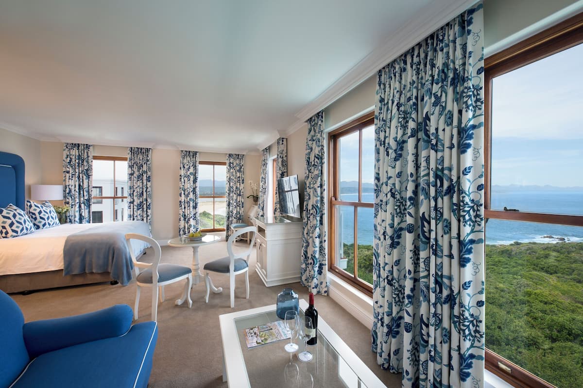 Une chambre d’hôtel avec un lit, un bureau, une chaise, une table avec une bouteille et des verres, et de grandes fenêtres donnant sur la mer.