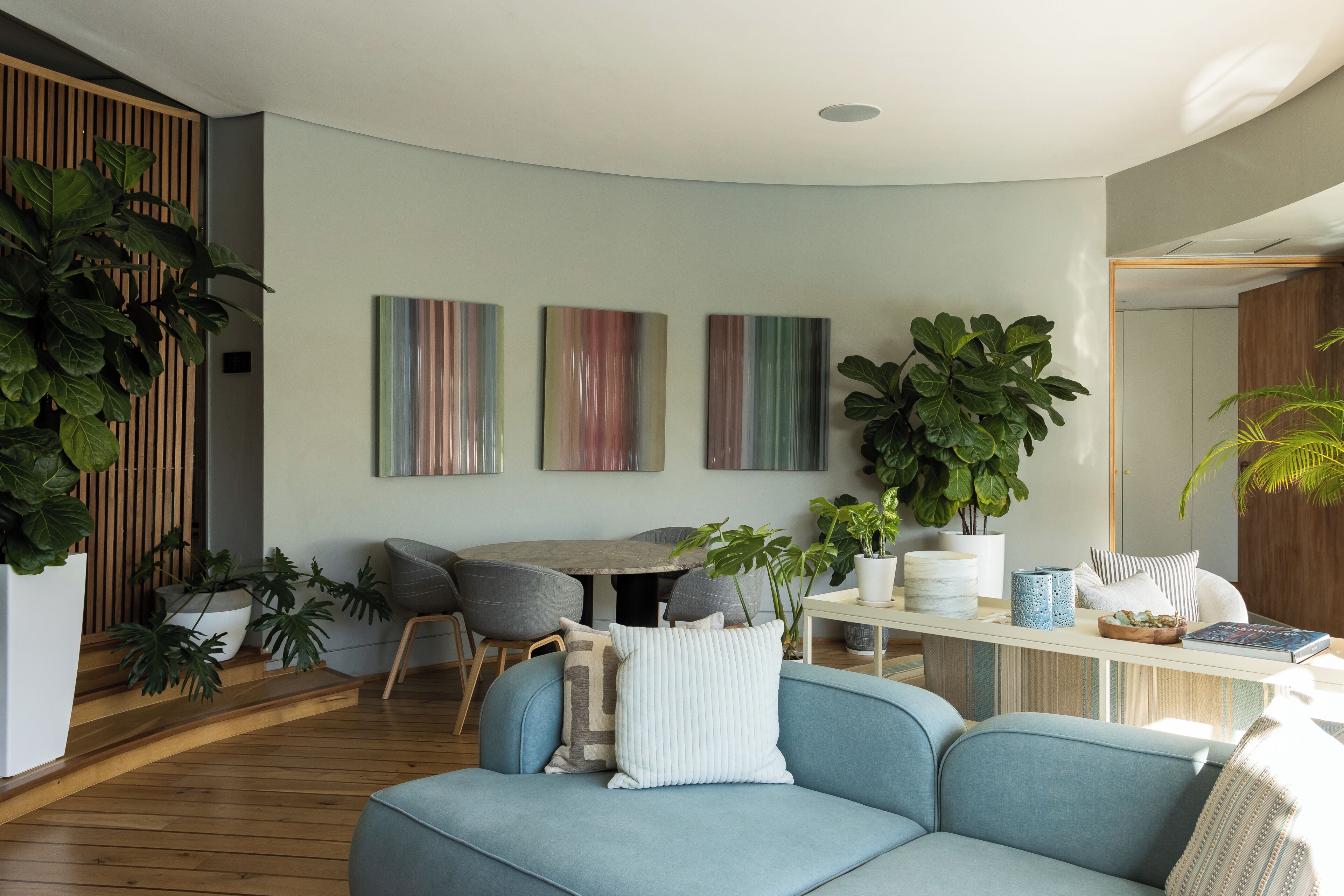 A modern living room with a blue sofa, wooden flooring, a dining table with chairs, and large green plants.