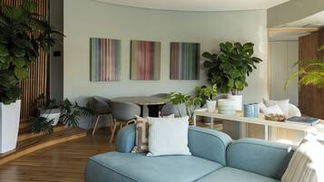 A modern living room with a blue sofa, wooden flooring, a dining table with chairs, and large green plants.