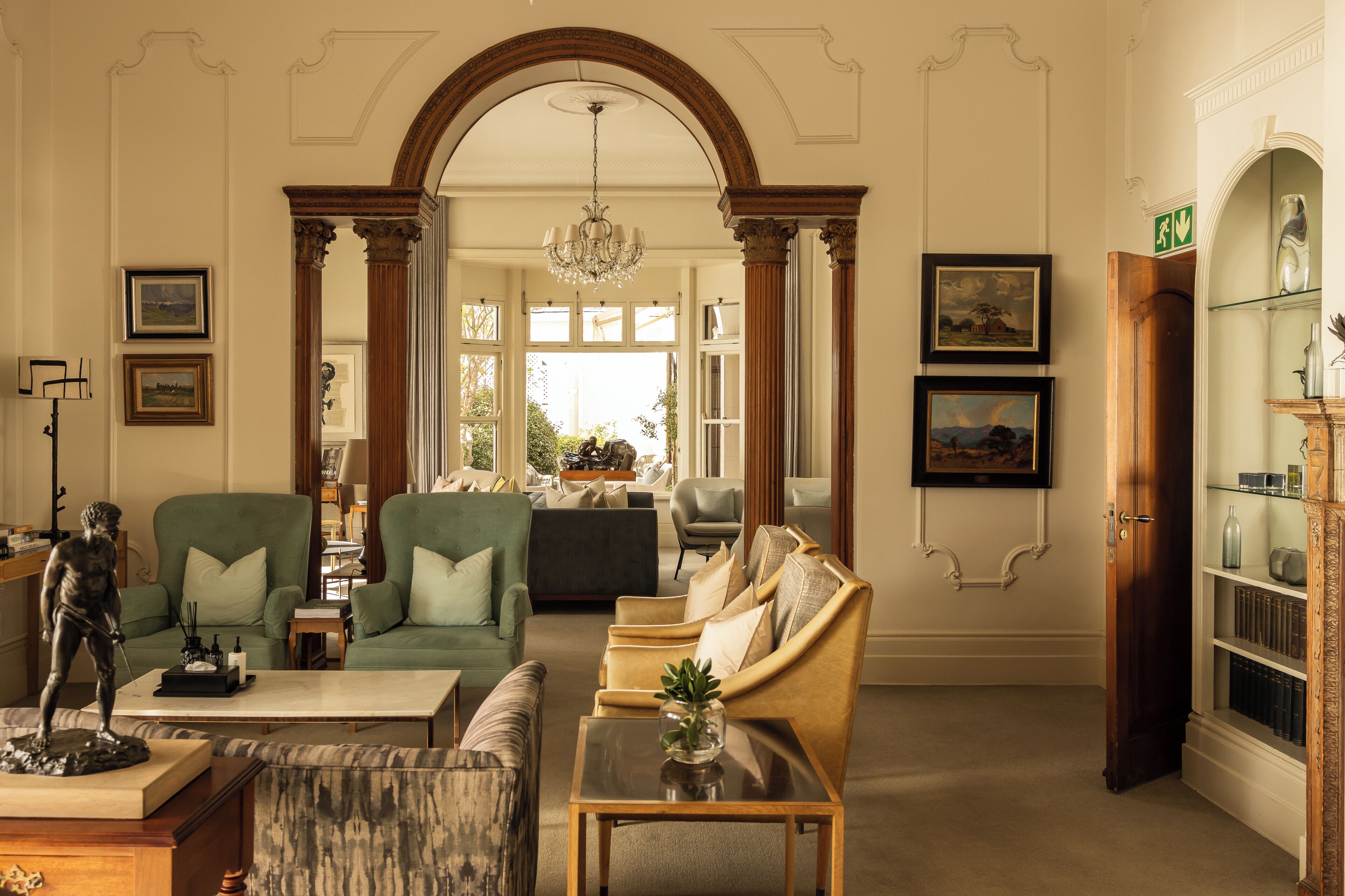 A spacious living room with a green sofa, a beige armchair, and a marble coffee table. There is a statue on a wooden stand, and a bookshelf with decorative items.