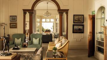 A spacious living room with a green sofa, a beige armchair, and a marble coffee table. There is a statue on a wooden stand, and a bookshelf with decorative items.