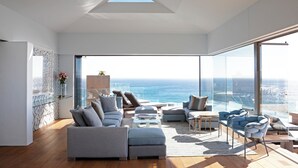 A modern living room with a large glass window overlooking the ocean, a sectional sofa, a coffee table, and a blue armchair. - Ellerman House (Cape Town)