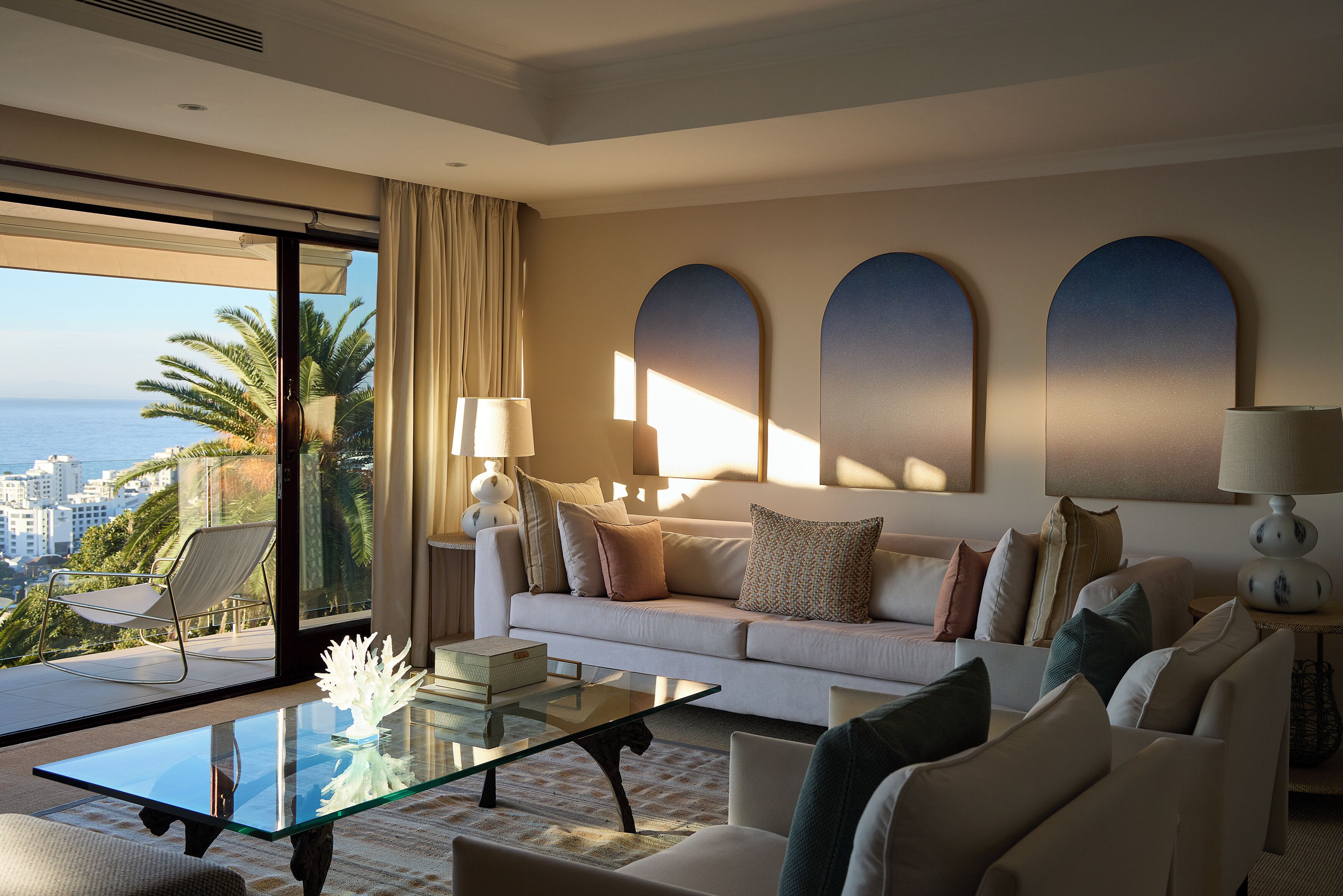 A modern living room with a glass coffee table, a sofa with multiple cushions, a lamp, and a view of the ocean.