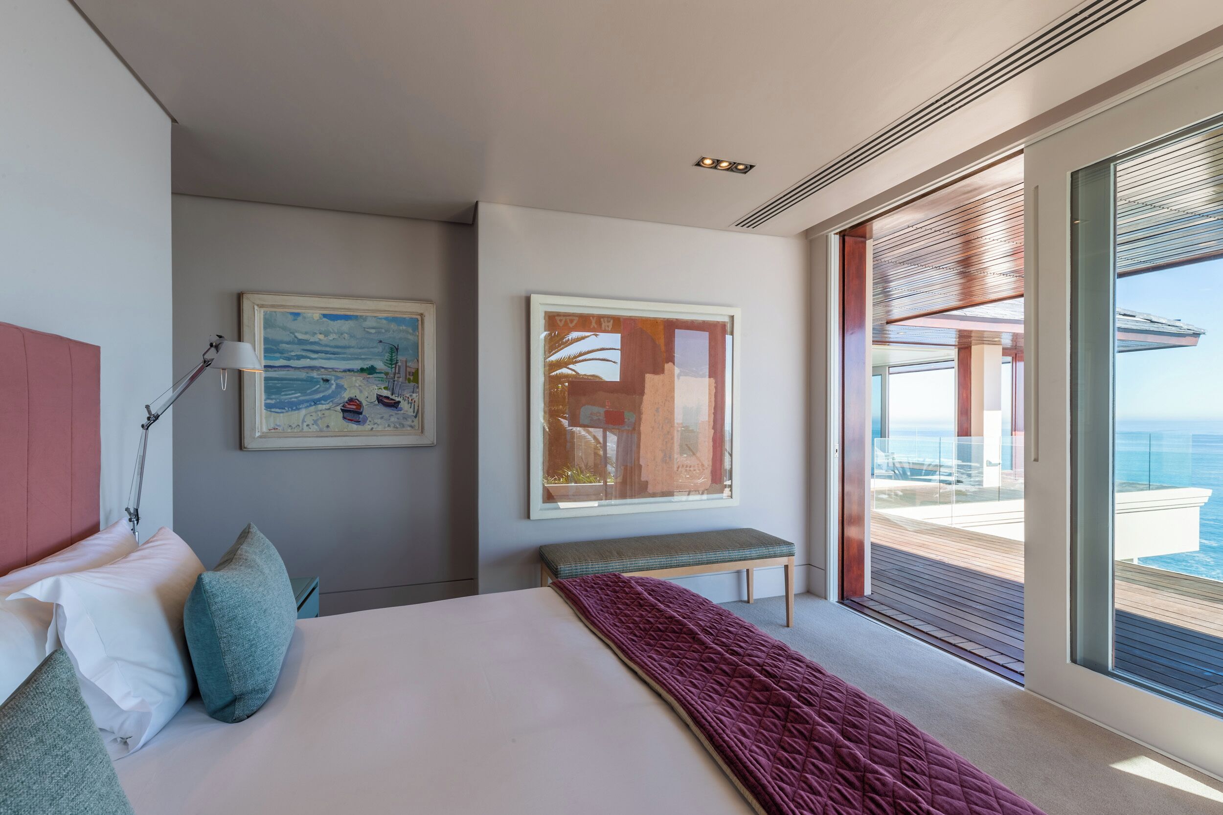 Un dormitorio moderno con una cama grande, dos cuadros, un banco y un balcón con vistas al mar.