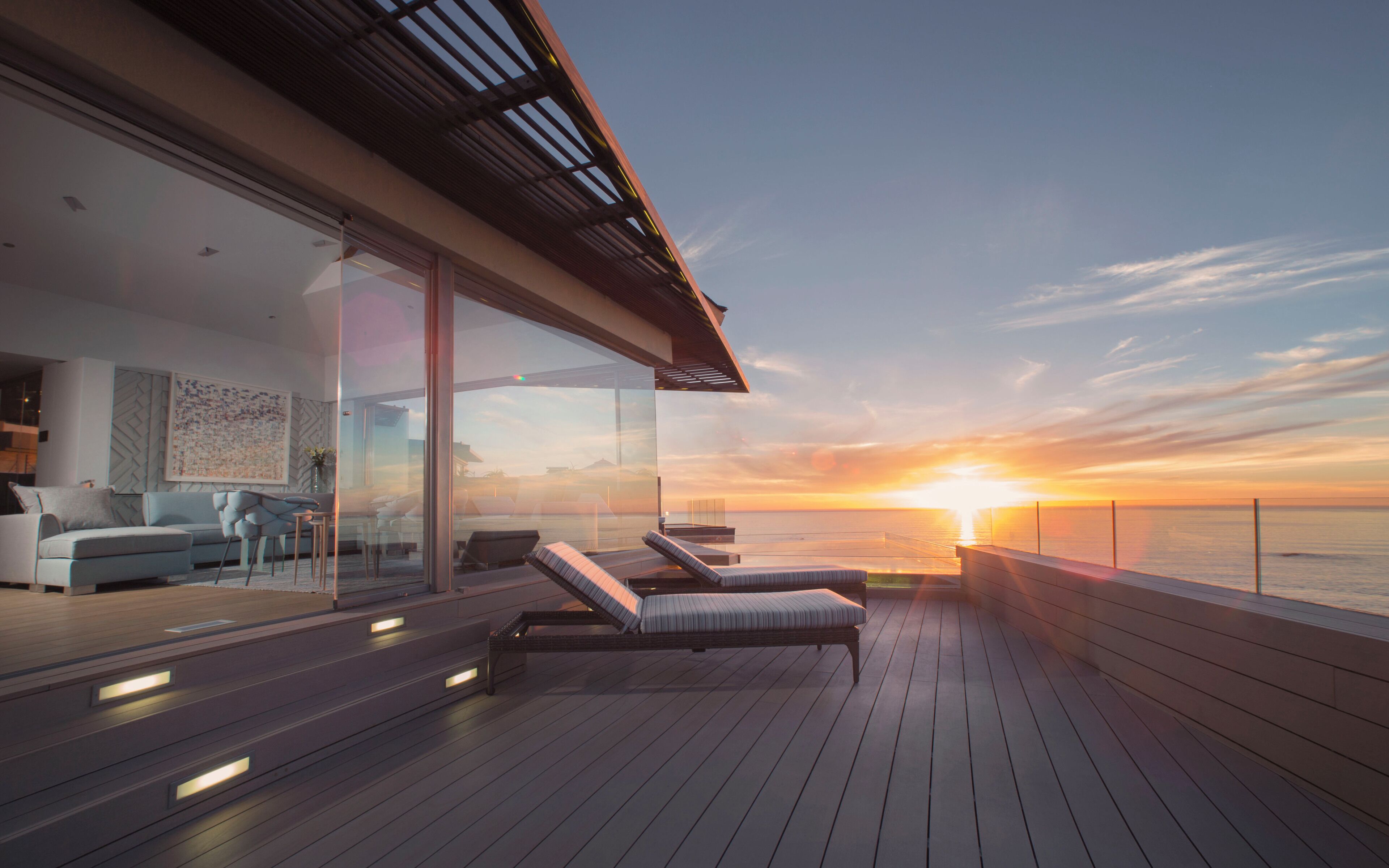A modern outdoor deck with lounge chairs and a glass wall overlooking the ocean at sunset.