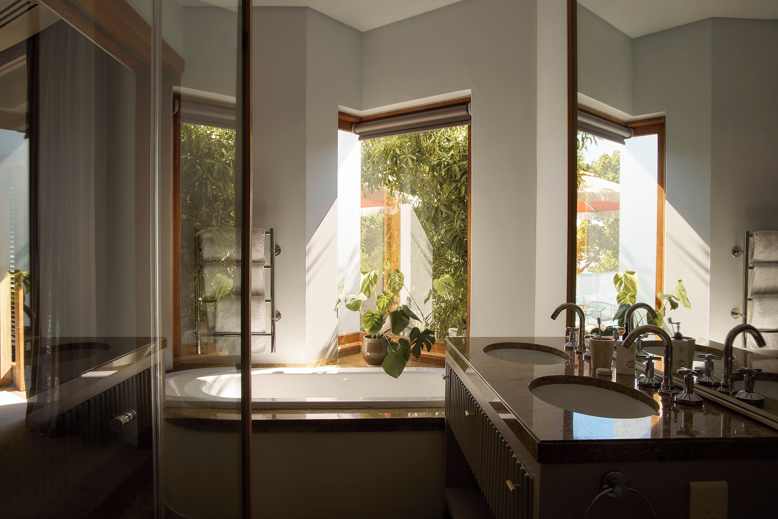 A bathroom with a bathtub, two sinks, a large mirror, and a view of greenery outside.