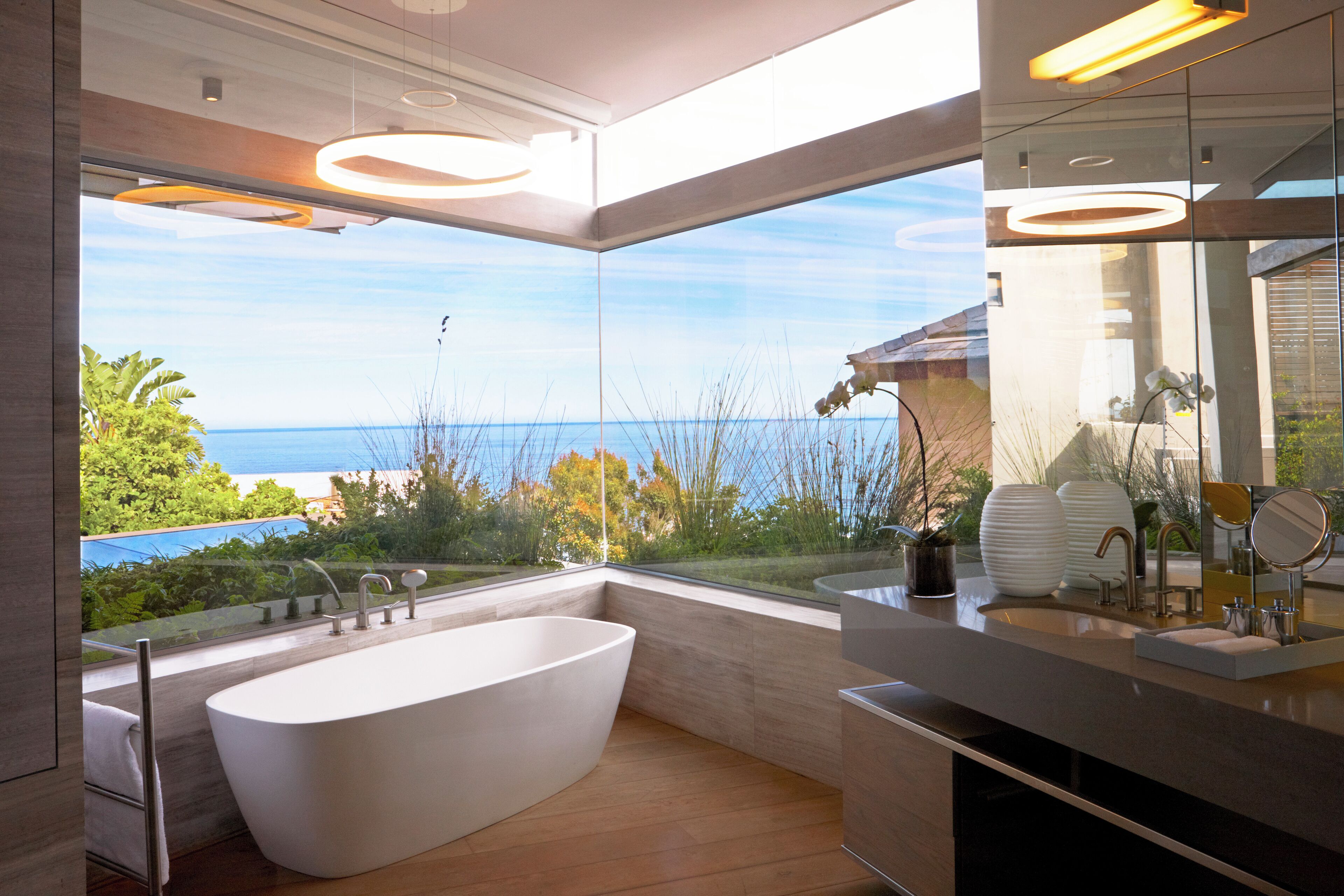A modern bathroom with a freestanding bathtub, a large mirror, and a view of the sea.