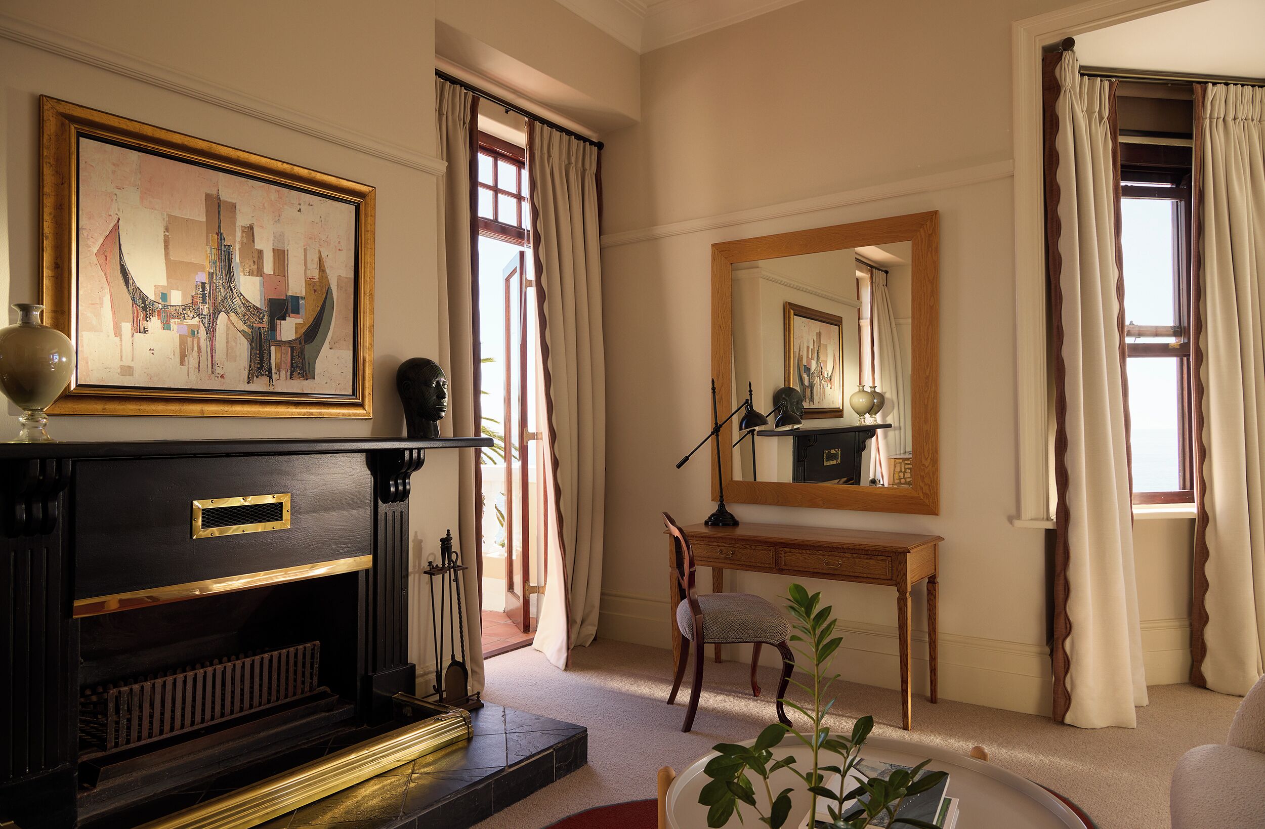 A room with a fireplace, a desk, a chair, a large mirror, and a painting on the wall.