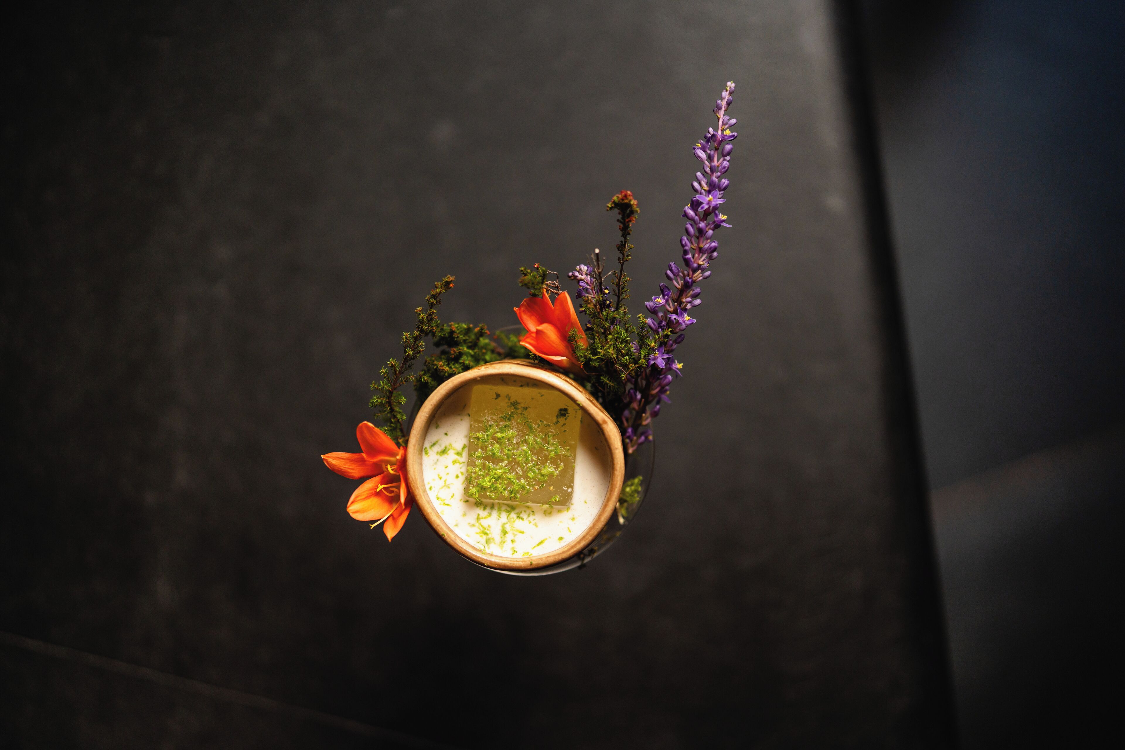 A top-down view of a small, round, ceramic bowl with a green substance, surrounded by various plants and flowers.
