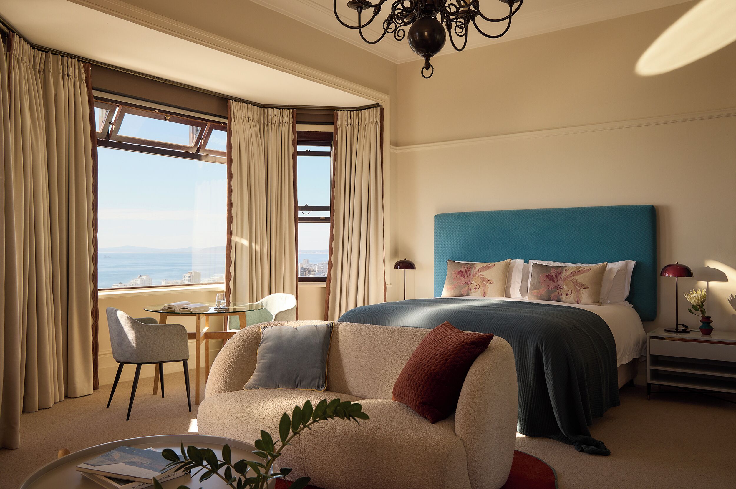 Habitación de hotel con cama, sofá, sillón, mesa y vista al océano.