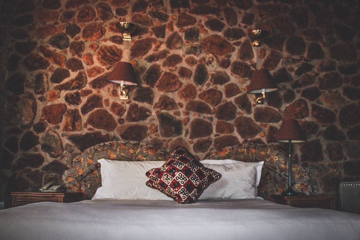 A bedroom with a stone wall, two bedside lamps, a patterned throw pillow, and a bedside table with a telephone.