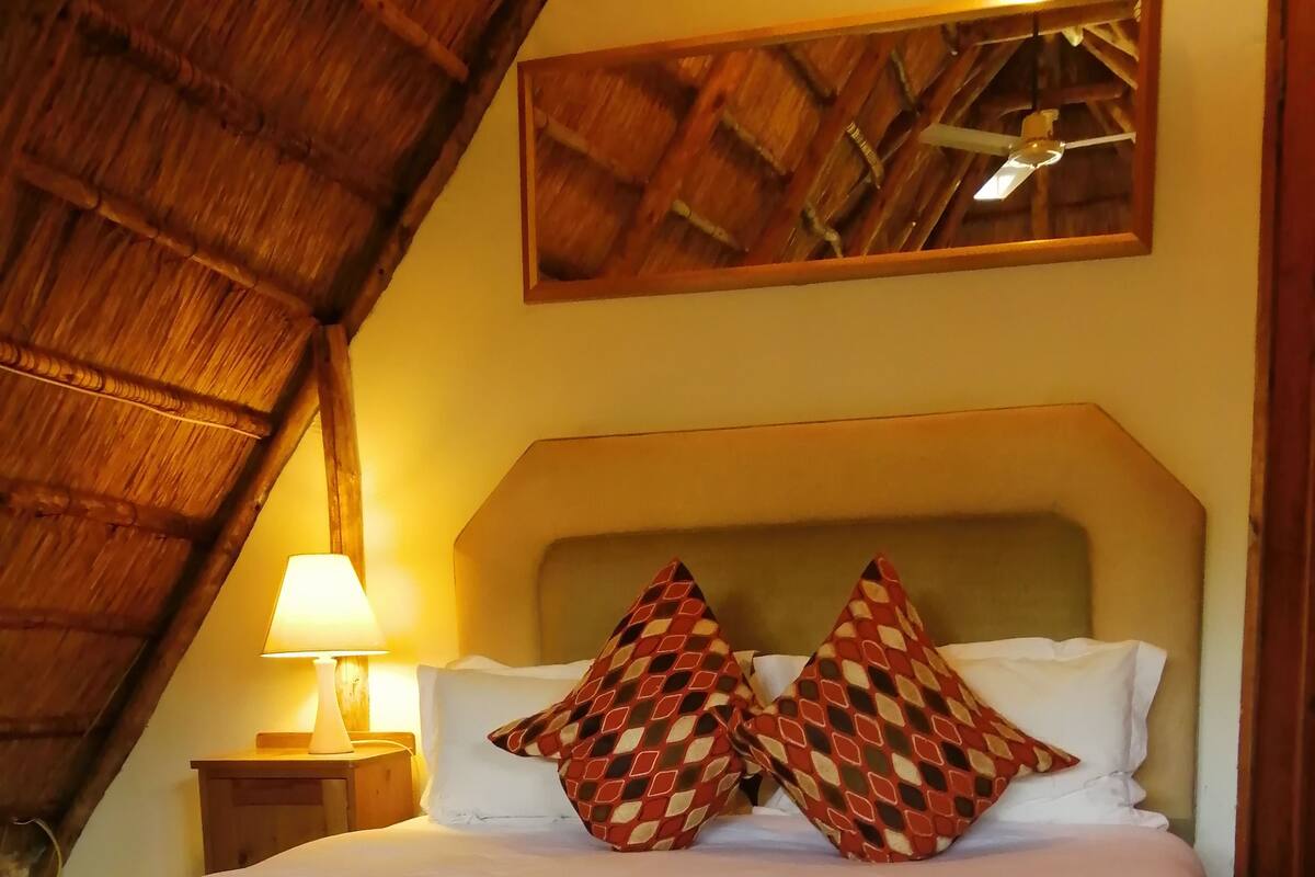 A bed with white linens and two patterned pillows, a nightstand with a lamp, and a wooden ceiling.