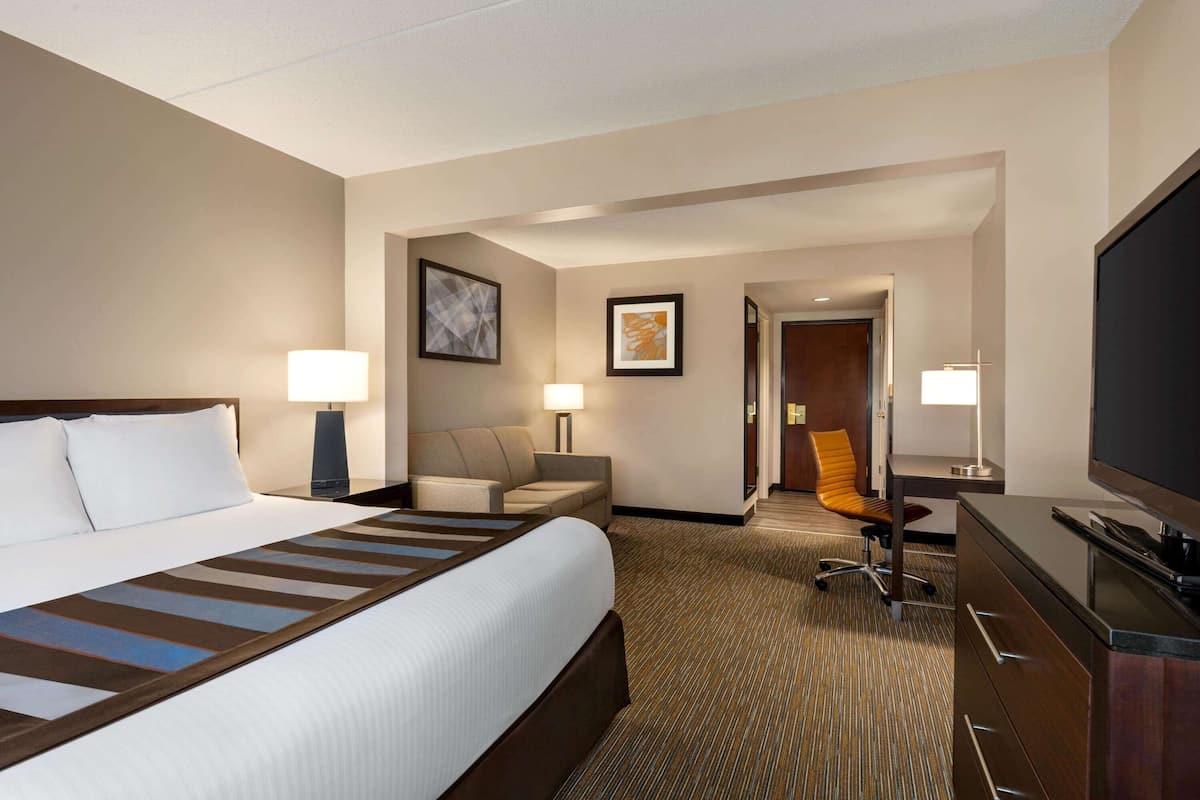 A hotel room with a large bed, a desk, a chair, a TV, and a door leading to another room.