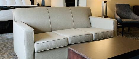 A hotel room with a sofa, a coffee table, a chair, a lamp, and a bed.