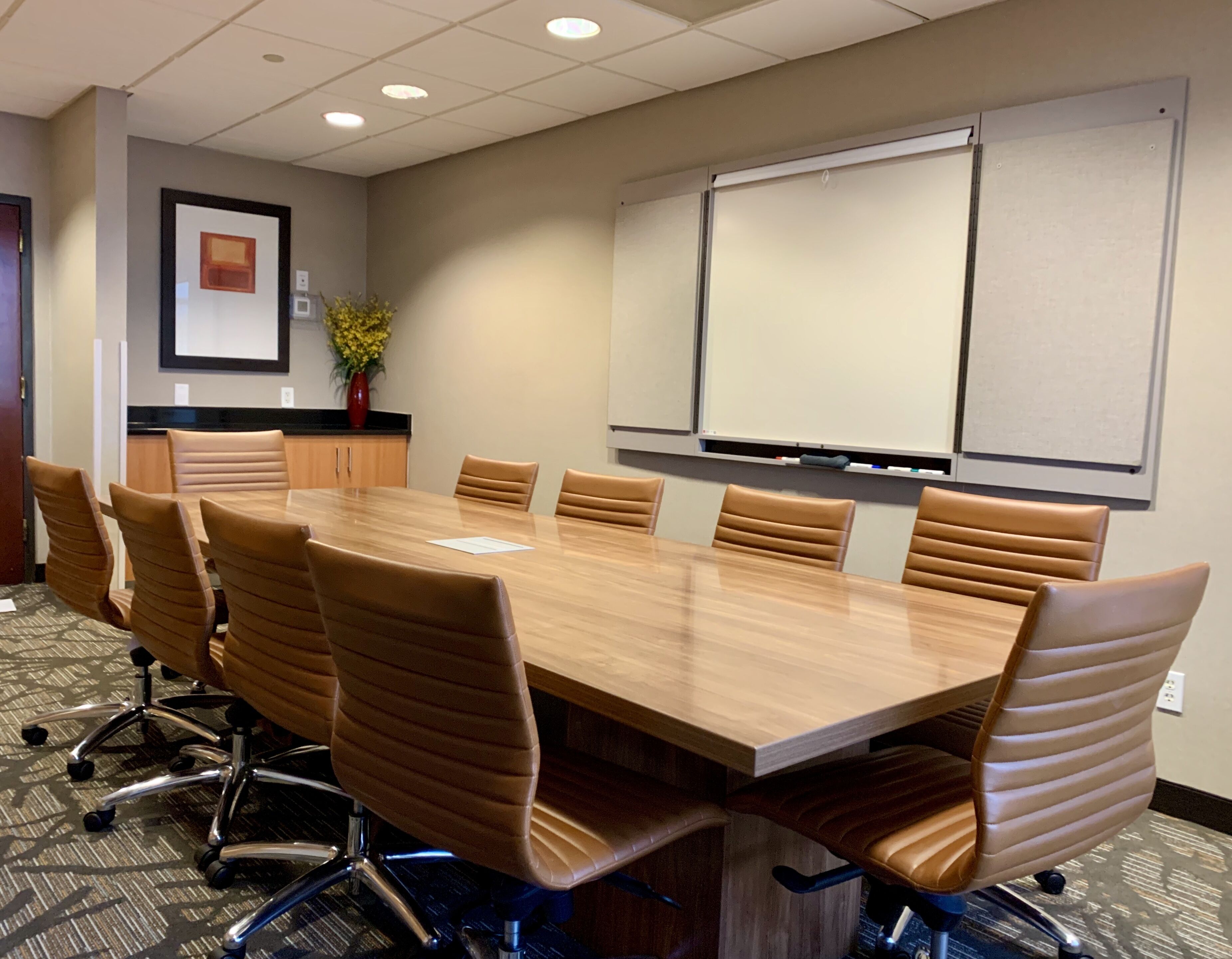 A conference room with a long table, six chairs, a whiteboard, and a framed picture on the wall.