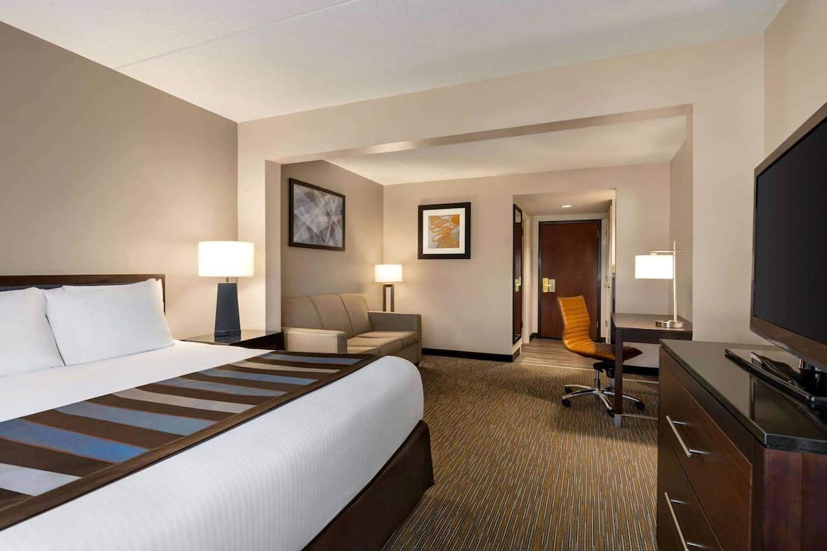 A hotel room with a large bed, a desk, a chair, a television, and a door leading to another room.
