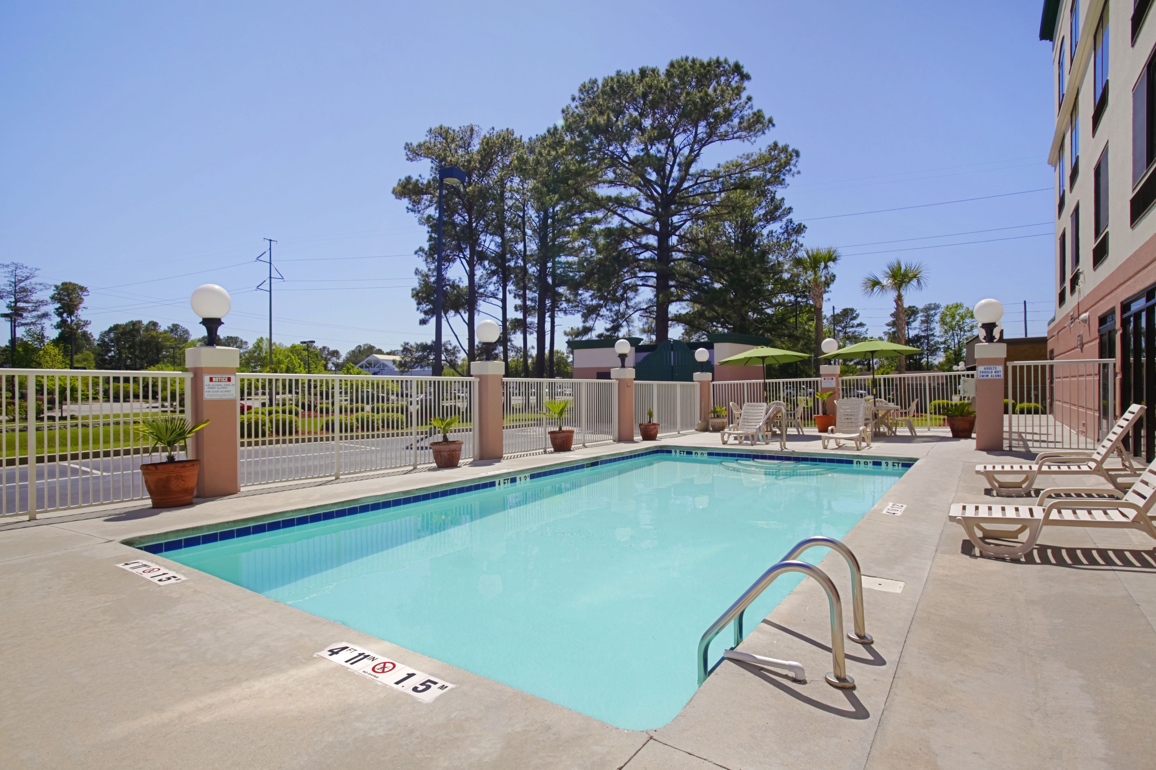 A rectangular swimming pool with a metal ladder, surrounded by a white fence. There are lounge chairs and potted plants along the poolside.