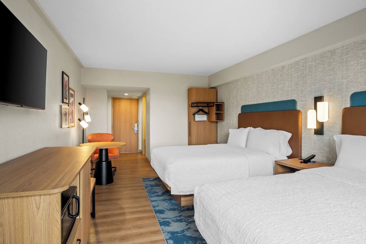 Une chambre d’hôtel avec deux lits, un bureau, une chaise, une télévision et une étagère fixée au mur.