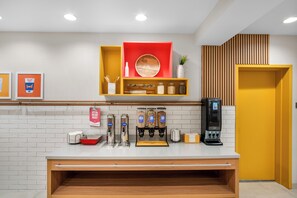 A modern hotel reception area with a white counter, a coffee machine, and a red shelf with various items.