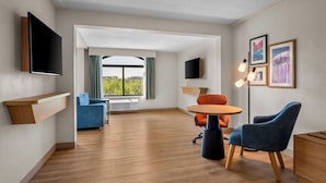 A modern hotel room with a flat-screen TV, a wooden shelf, a blue sofa, an orange chair, a round table, and a desk with drawers. - Hampton Inn by Hilton Addison Dallas (Addison)