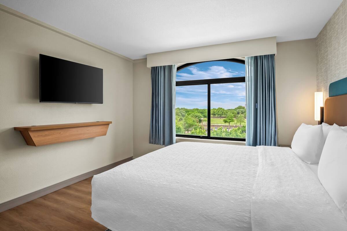 Une chambre d’hôtel avec un grand lit, un téléviseur fixé au mur, une fenêtre avec des rideaux et une étagère en bois.