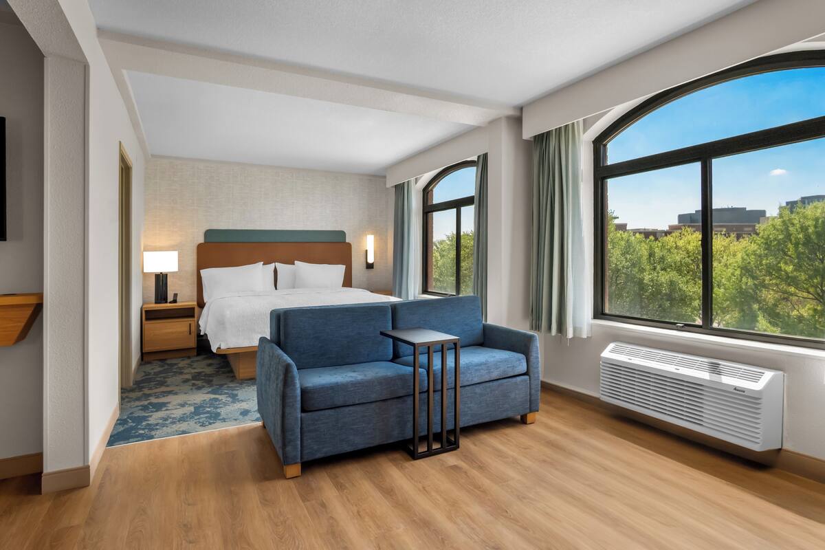 Une chambre d’hôtel avec un lit, un canapé bleu, une table de chevet, une lampe, une fenêtre donnant sur un beau paysage et un climatiseur fixé au mur.