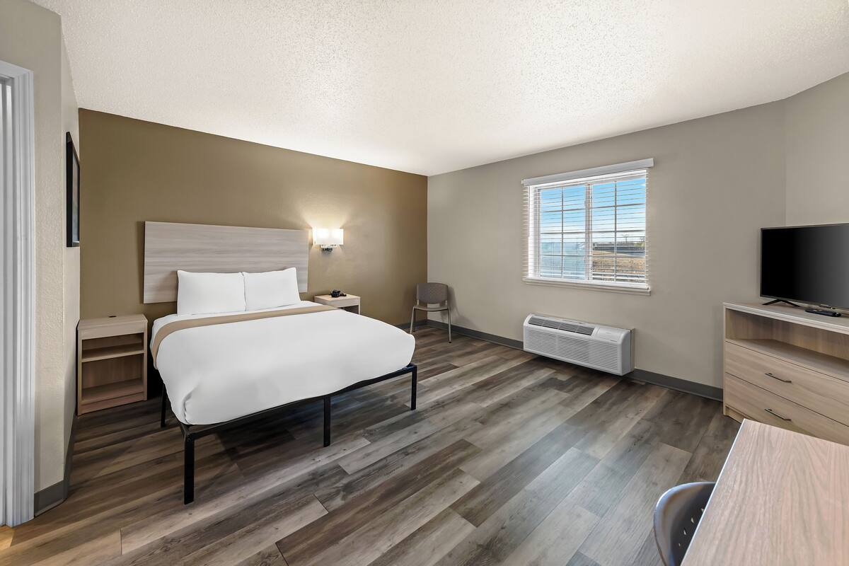 Habitación de hotel con cama, mesita de noche, lámpara, televisor y ventana con persianas.