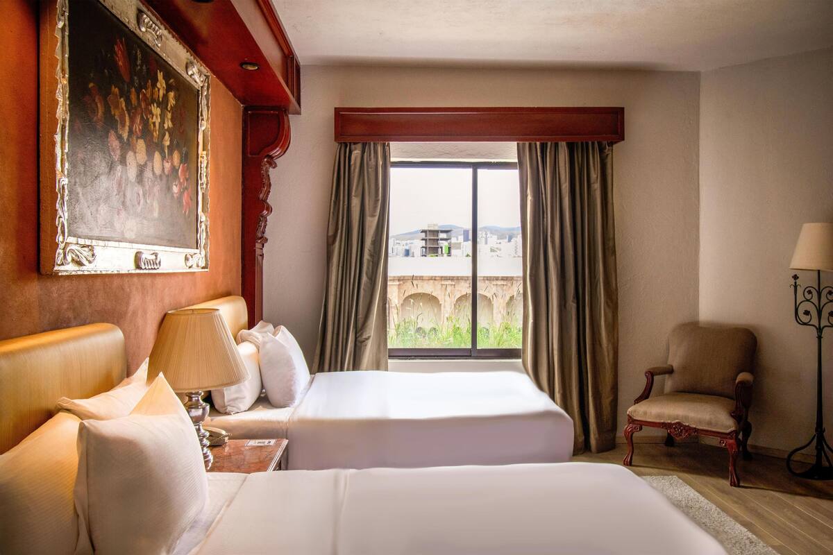 A hotel room with two beds, a painting, a view of a cityscape, and a chair.