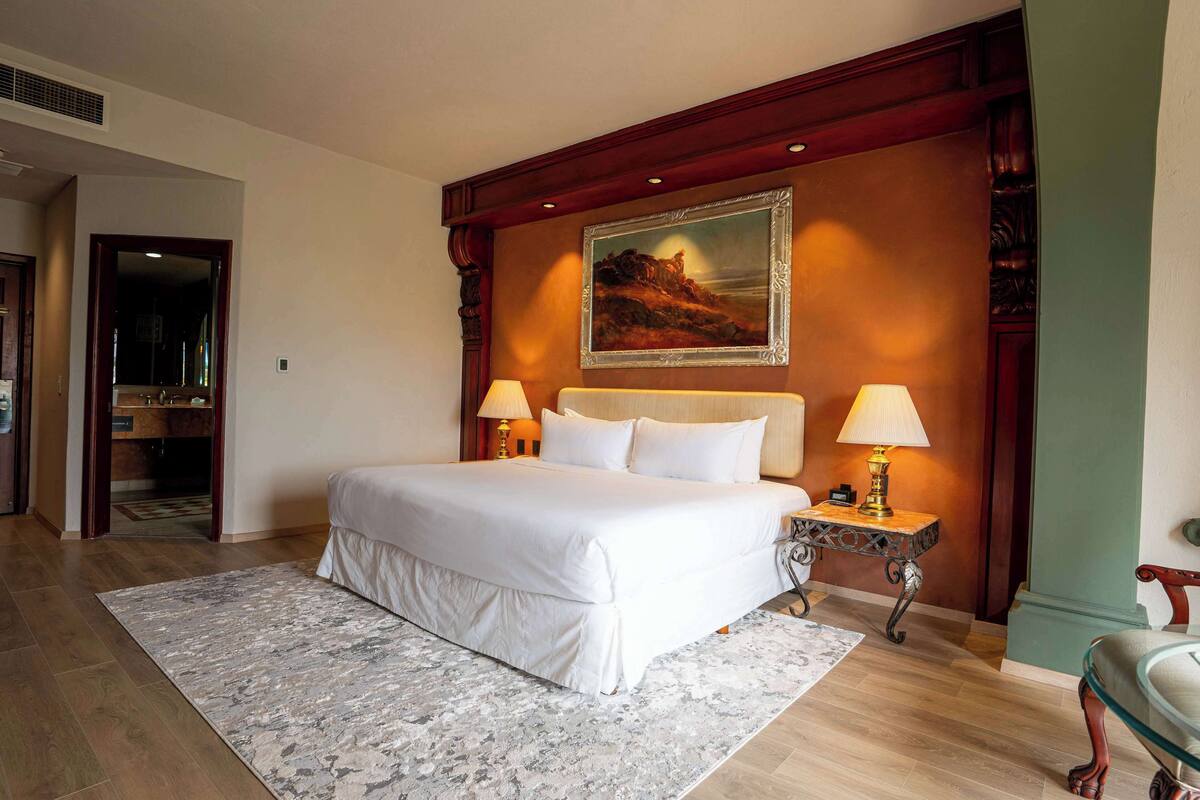 A hotel room with a large bed, two bedside lamps, a framed painting on the wall, and a wooden headboard.