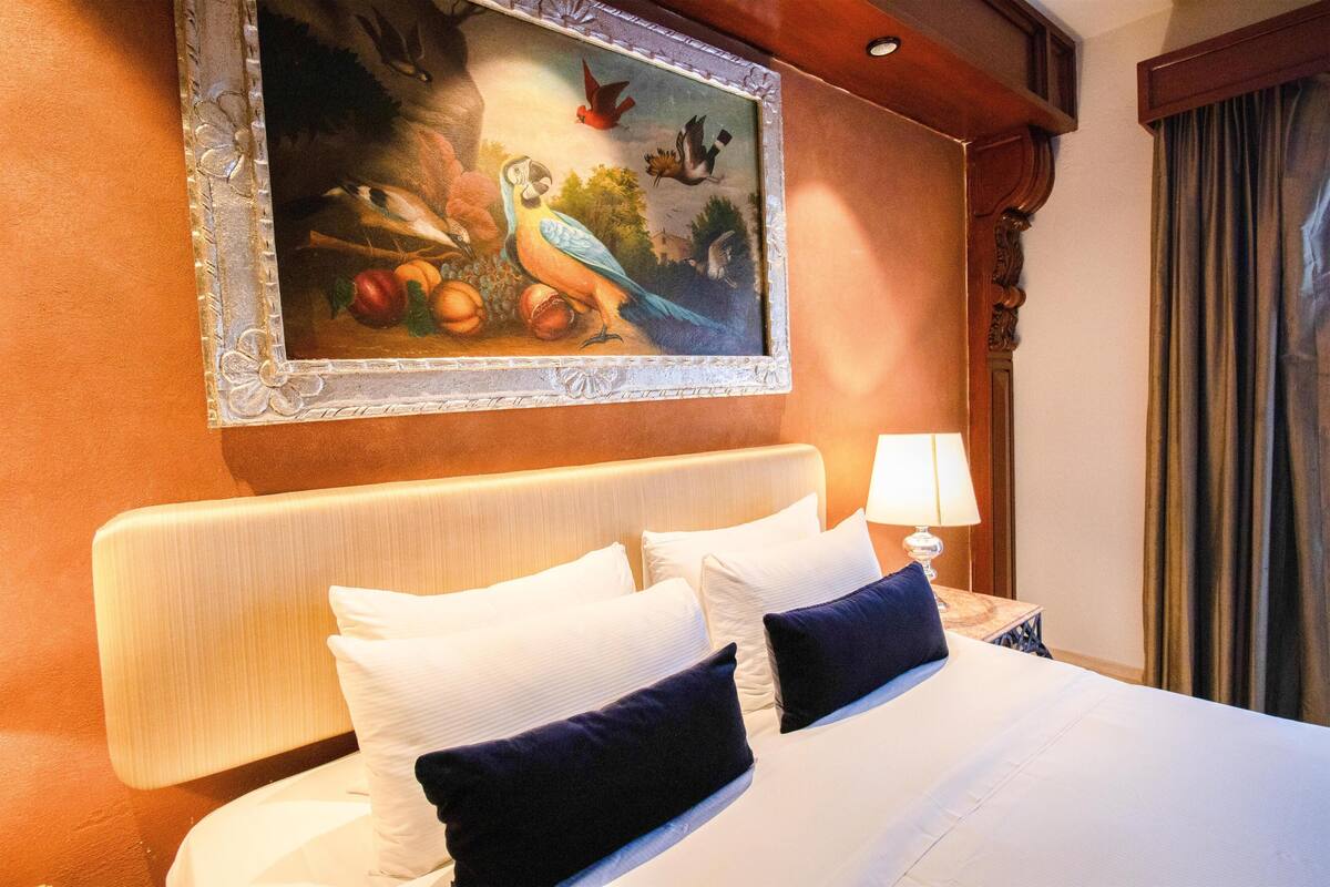 A hotel room with a large bed, white bedding, a headboard, a nightstand with a lamp, and a painting on the wall.