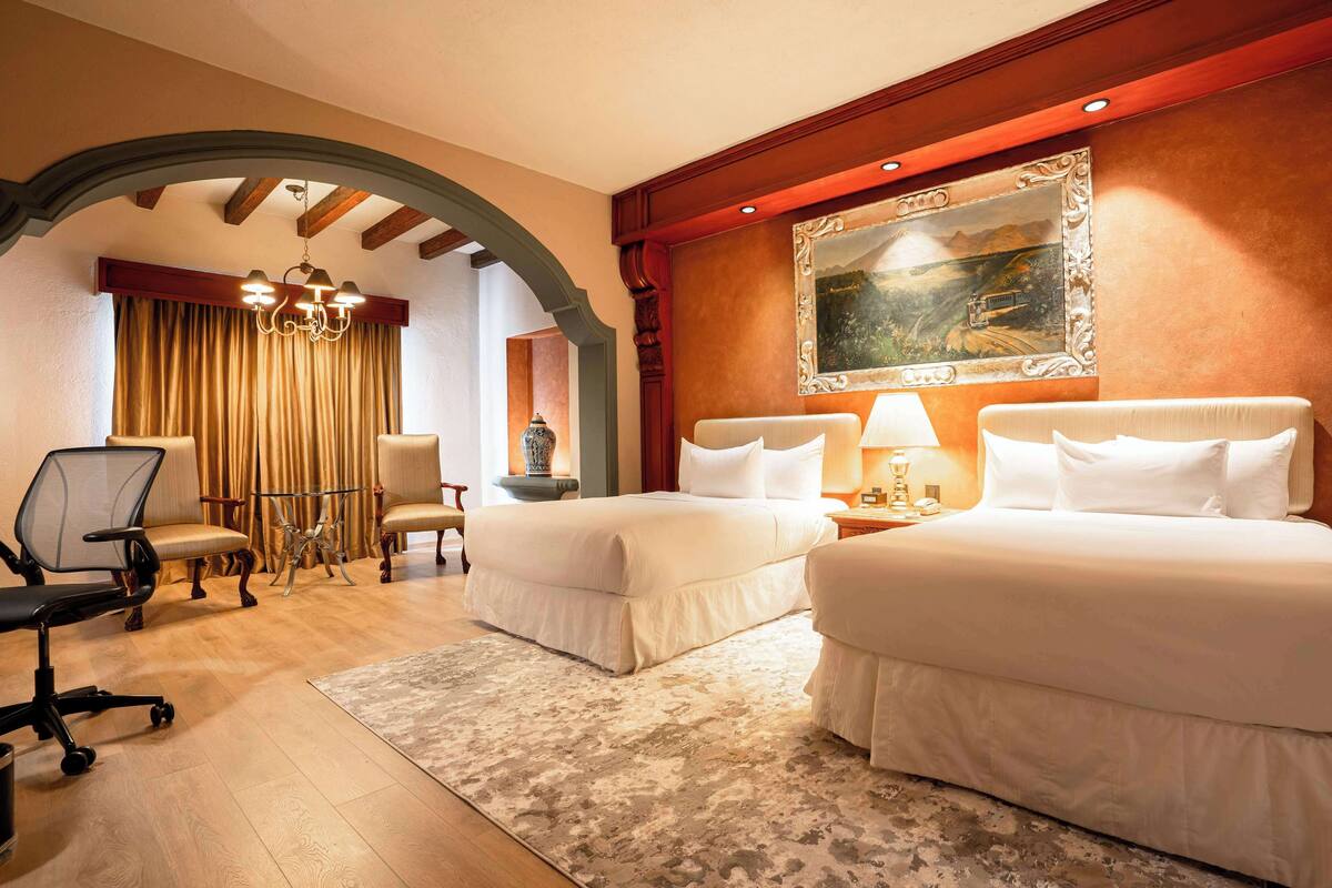 A hotel room with two beds, a desk with a chair, a chandelier, and a large painting on the wall.
