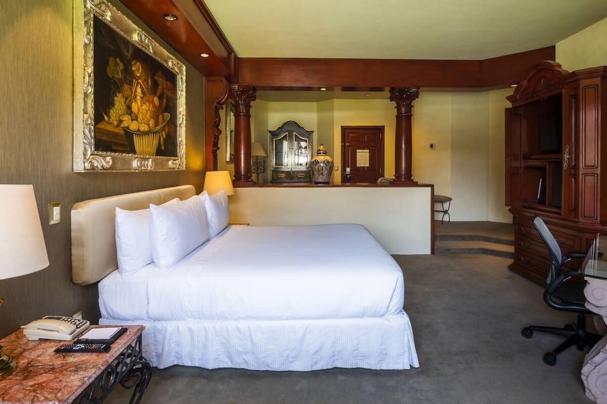 A hotel room with a large bed, a desk with a telephone and a laptop, a chair, and a mirror.