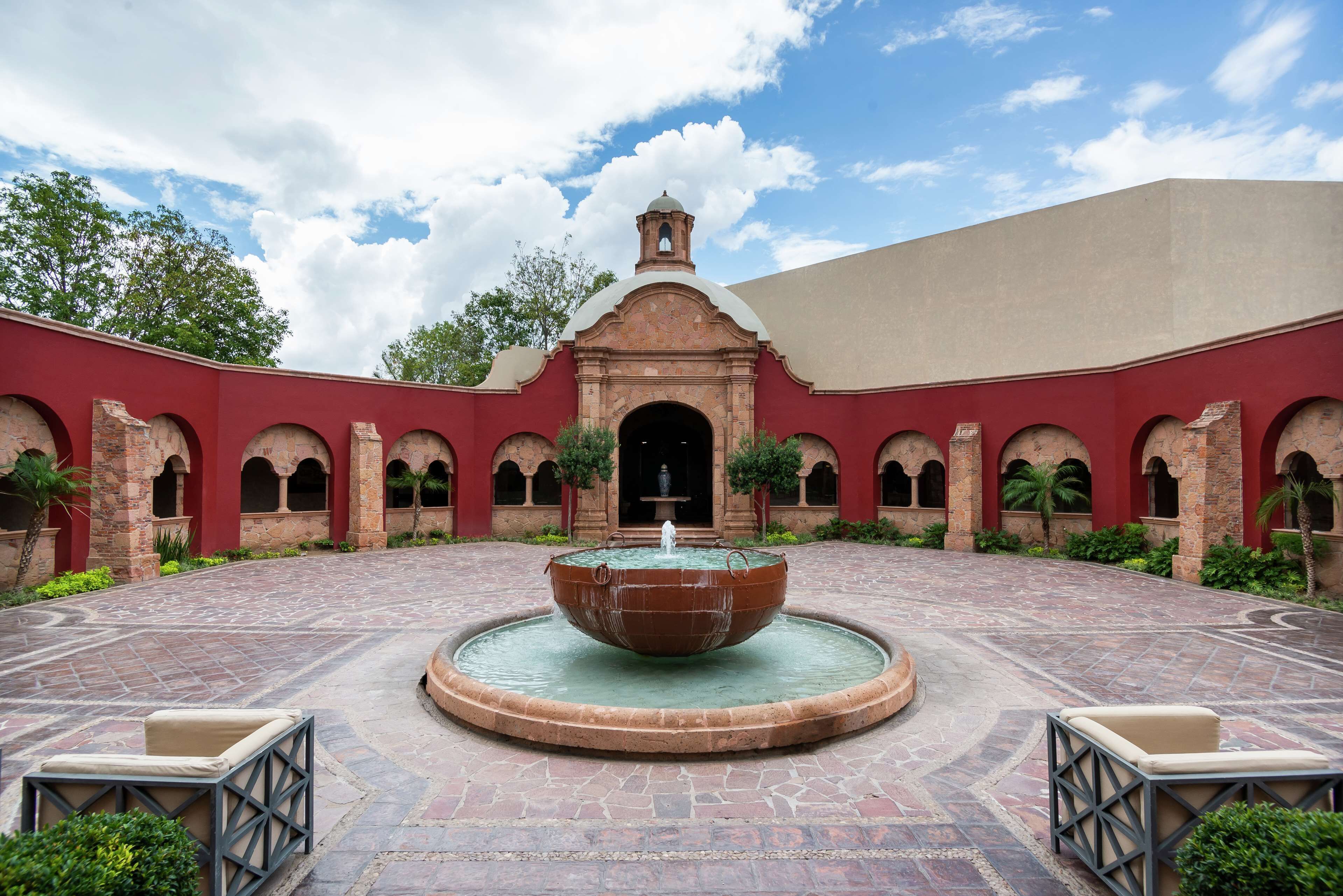 A courtyard with a central fountain, surrounded by red brick arches and a building with a dome and bell tower.