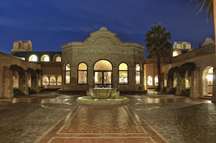 Un patio con una fuente, arcos de piedra y un edificio con ventanas arqueadas.