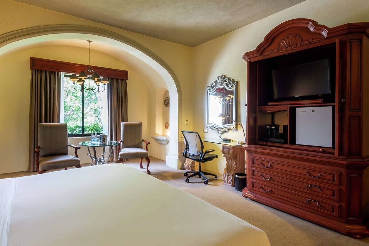 A hotel room with a large bed, two armchairs, a desk with a computer, a television, and a mirror.