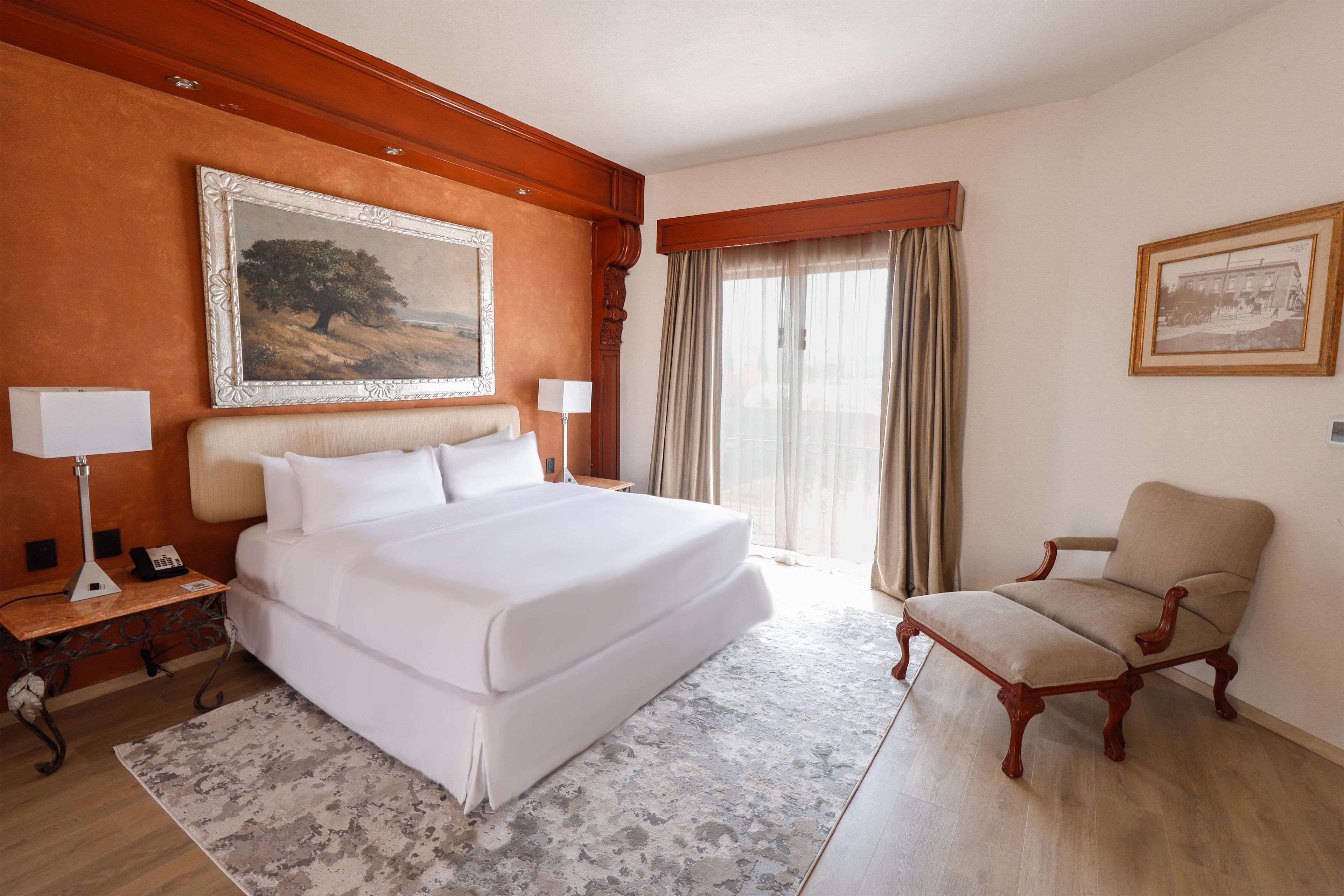 A hotel room with a large bed, a desk with a lamp, a chair, and two framed pictures on the walls.