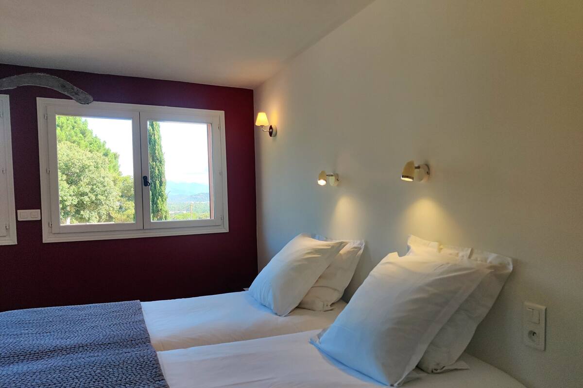 Une chambre avec deux lits, une fenêtre donnant sur un espace verdoyant et des lampes fixées au mur.