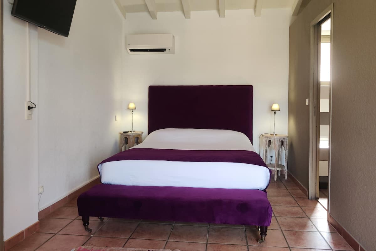 Une chambre à coucher comprenant un lit, deux tables de chevet, un téléviseur à écran plat, un appareil de climatisation et une porte donnant sur une autre pièce.