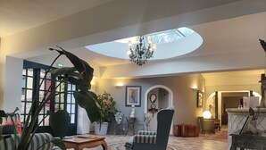 Een ruime lobby met een hoge plafond, een dakvenster, een kroonluchter, een grote potplant, een gestreepte bank, een houten salontafel en een receptiebalie.