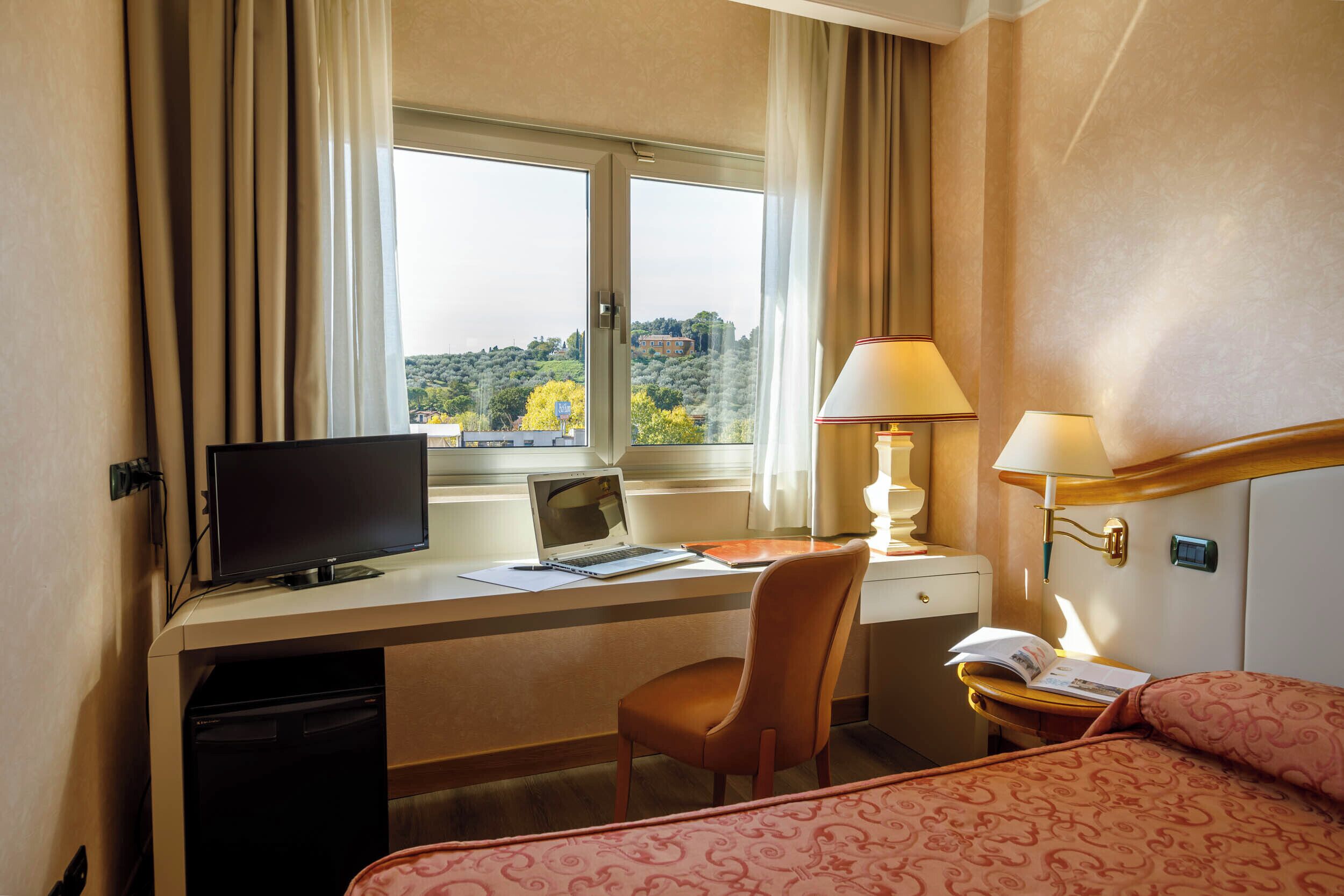 Habitación de hotel con escritorio, laptop, silla, lámpara de noche y cama con ropa de cama estampada.