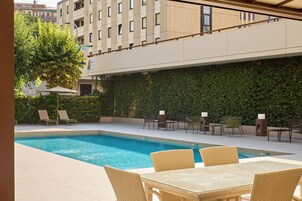 Piscina con sillones y mesas, rodeada por un edificio con pared verde y cielo despejado.