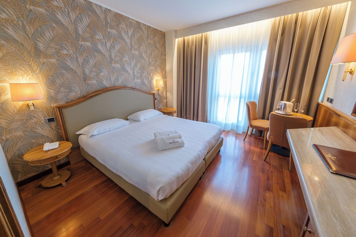 A hotel room with a large bed, a desk, a chair, a lamp, and a window with curtains.