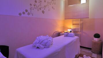 A spa room with a massage table, towels, a small plant, and a window with blinds.