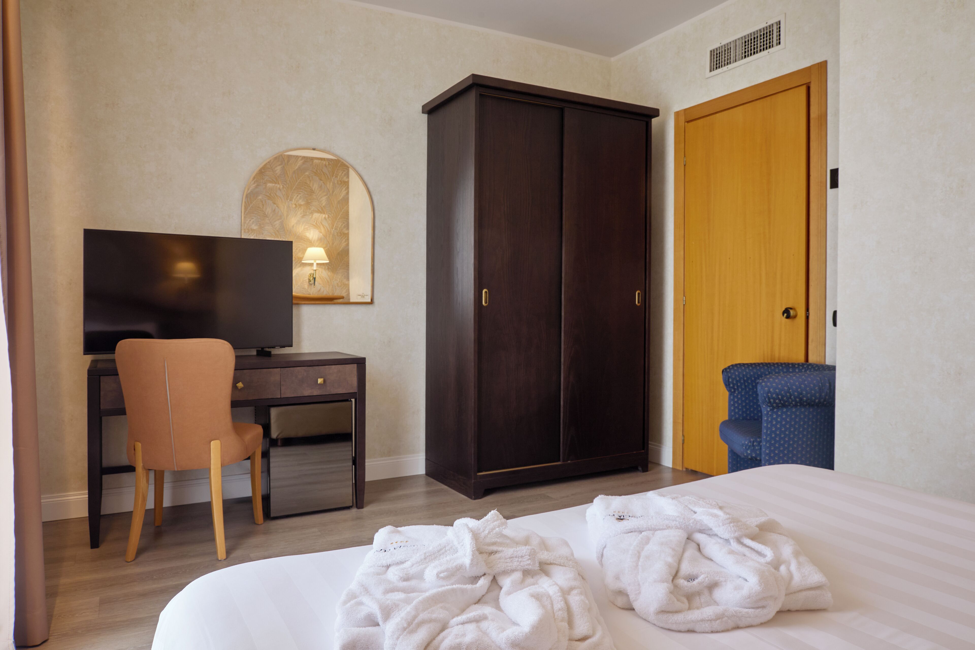 A hotel room with a bed, a desk with a chair, a wardrobe, a television, and a blue armchair.