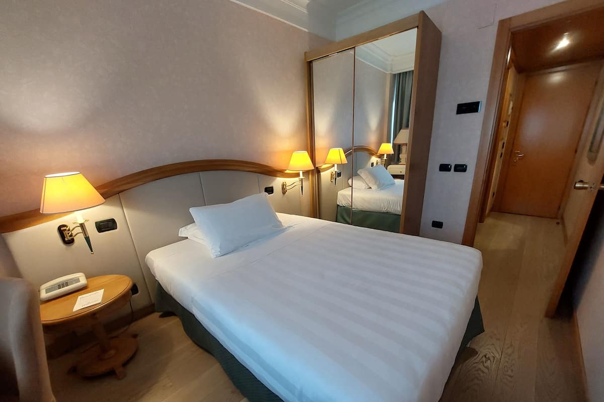 A hotel room with a bed, bedside lamps, a telephone, a mirror, and a door.