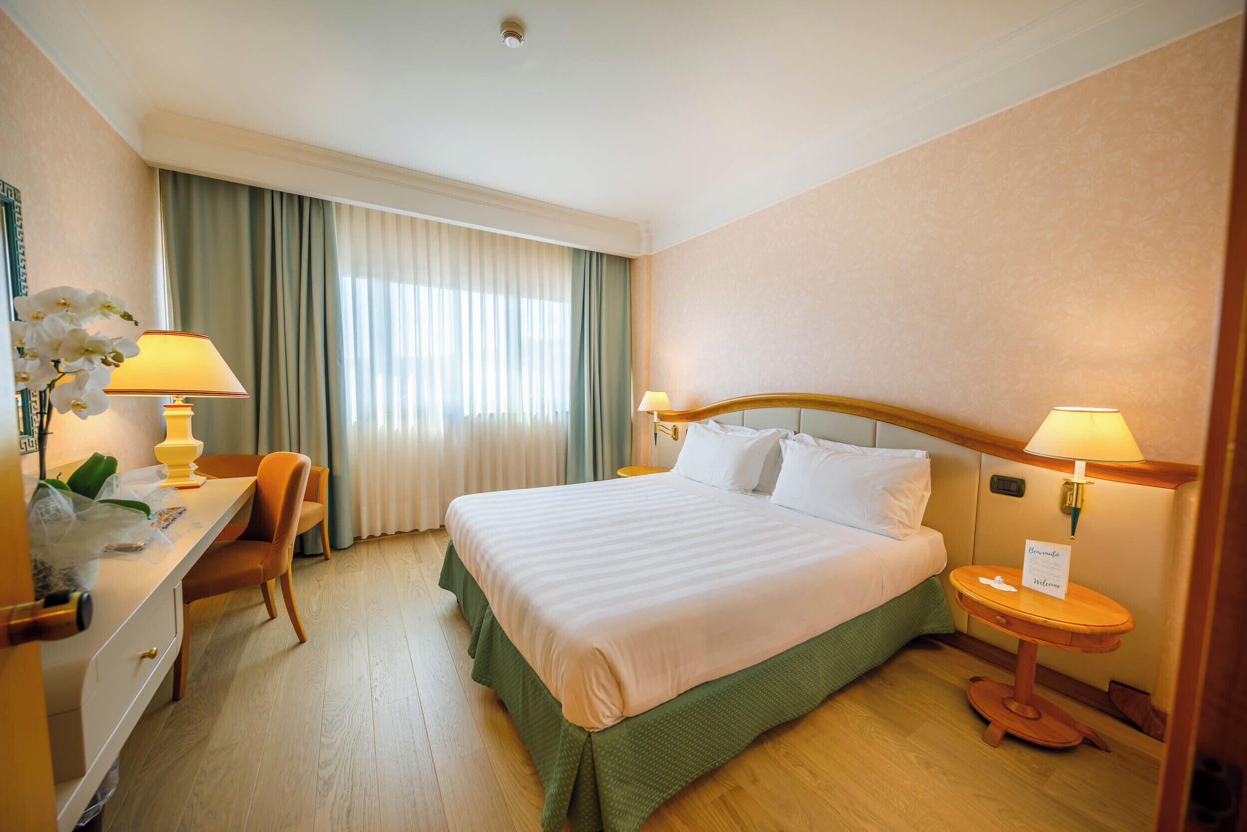 Une chambre d’hôtel avec un lit, deux lampes de chevet, un bureau avec une chaise et une fenêtre avec des rideaux.