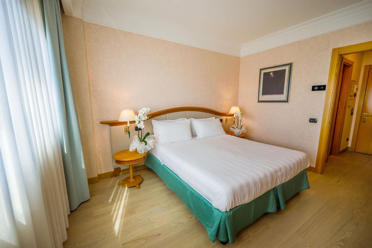 A hotel room with a large bed, two bedside lamps, a small table with a flower, and a door to the right.