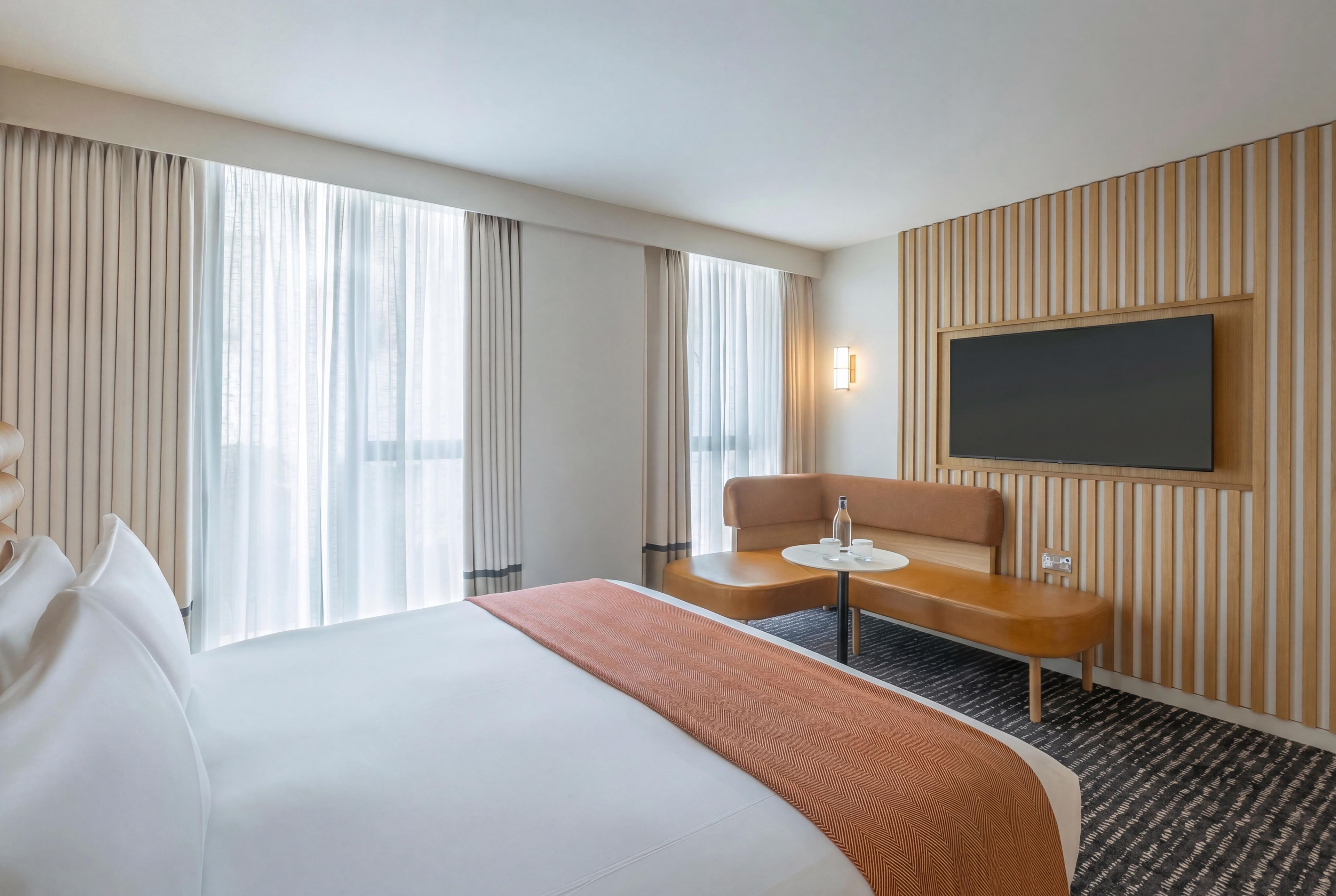 A hotel room with a large bed, a small table with a chair, a flat-screen TV mounted on the wall, and a window with curtains.
