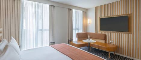 A hotel room with a large bed, a small table with a chair, a flat-screen TV mounted on the wall, and a window with curtains.