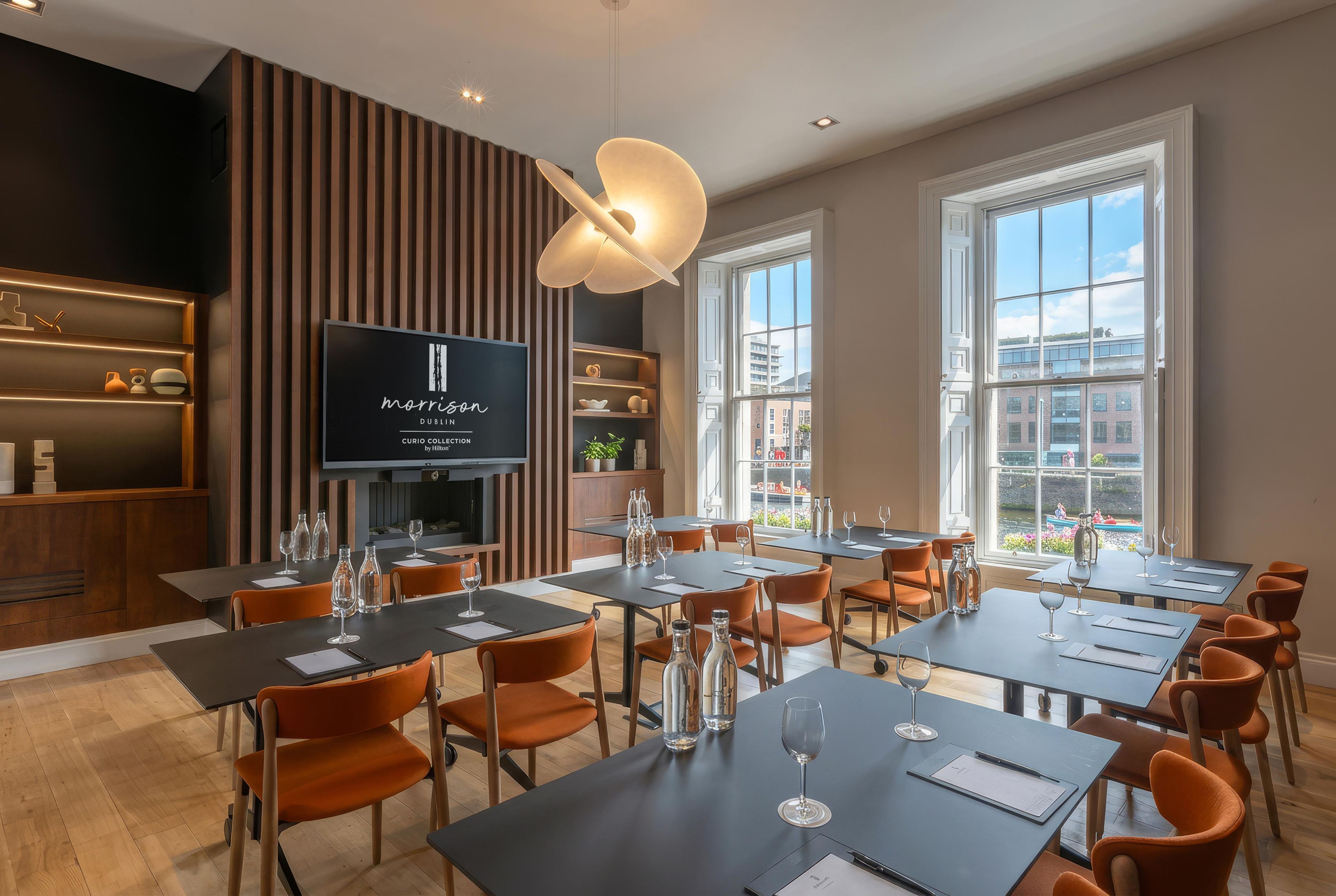 A modern hotel conference room with tables, chairs, a television screen, and large windows offering city views.